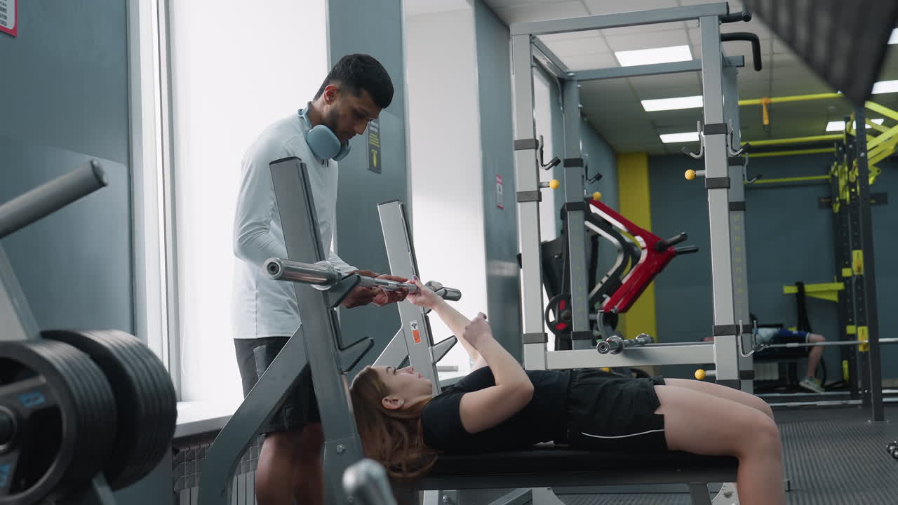 young woman lies on bench lifting barbell under guidance of male instructor in modern gym with weightlifting equipment and racks visible in background