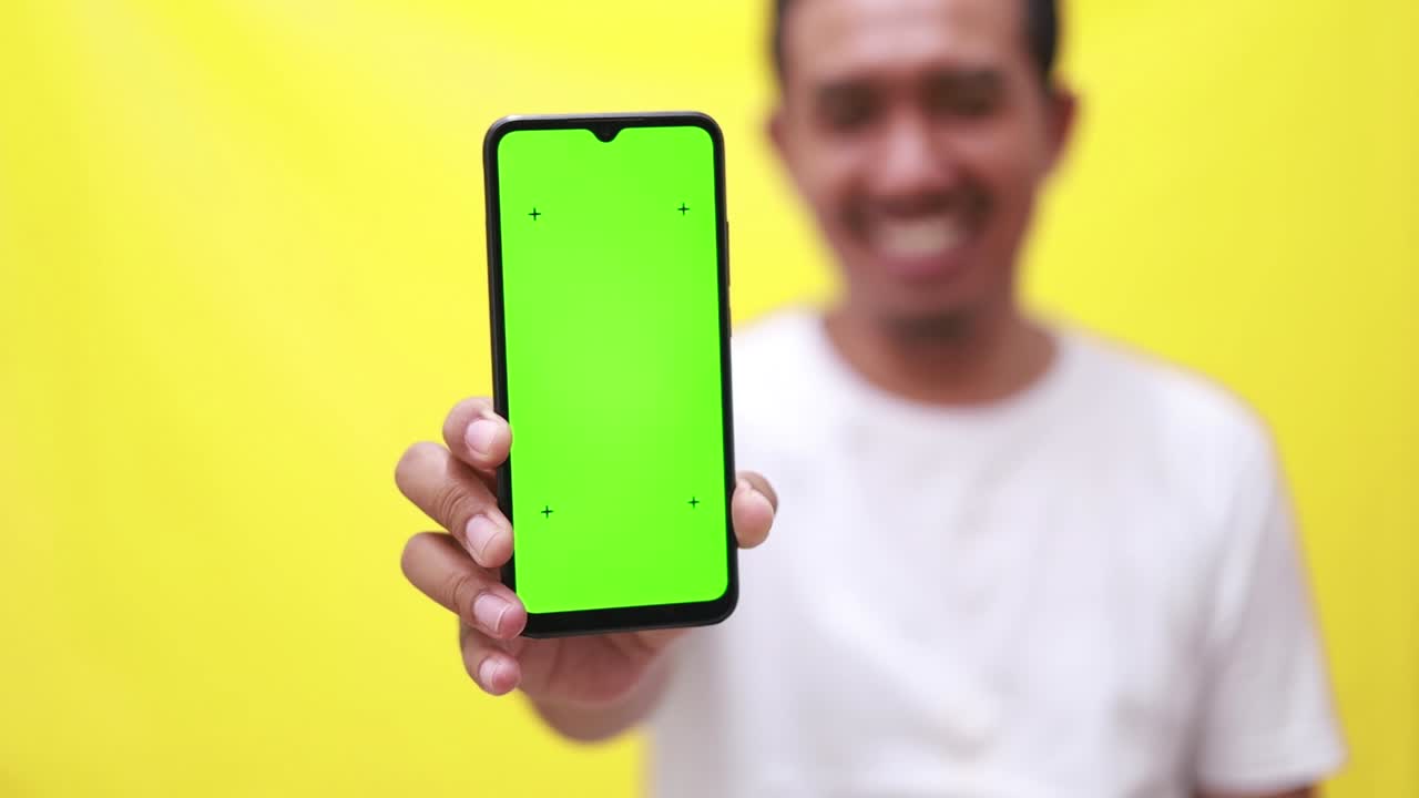 hombre adulto asiático presentando pantalla verde en la pantalla del teléfono celular mientras señala y muestra los pulgares hacia arriba