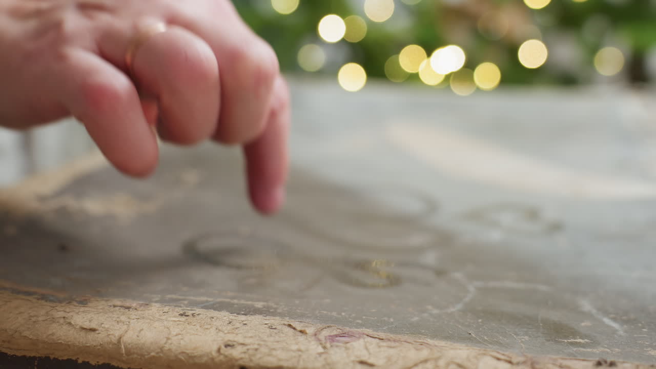 Hand view sketching shapes on dusty box surface with finger, capturing tactile creative moment during festive indoor setting with blurred glowing Christmas tree lights in background