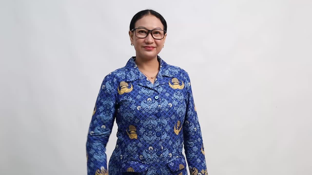 A cheerful Indonesian woman in blue Korpri batik uniform smiles confidently while making an "OK" hand gesture against a plain white background