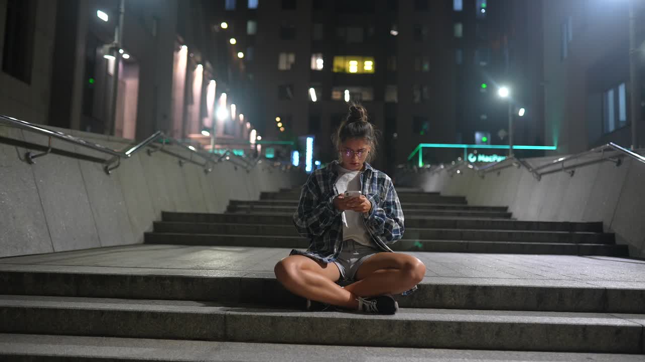 mujer joven sentada en las escaleras de la noche, usando un teléfono inteligente