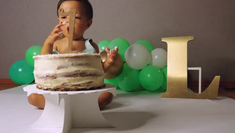 cute latin baby toddler celebrating his 1st year birthday smashing a cake with green balloons in the background