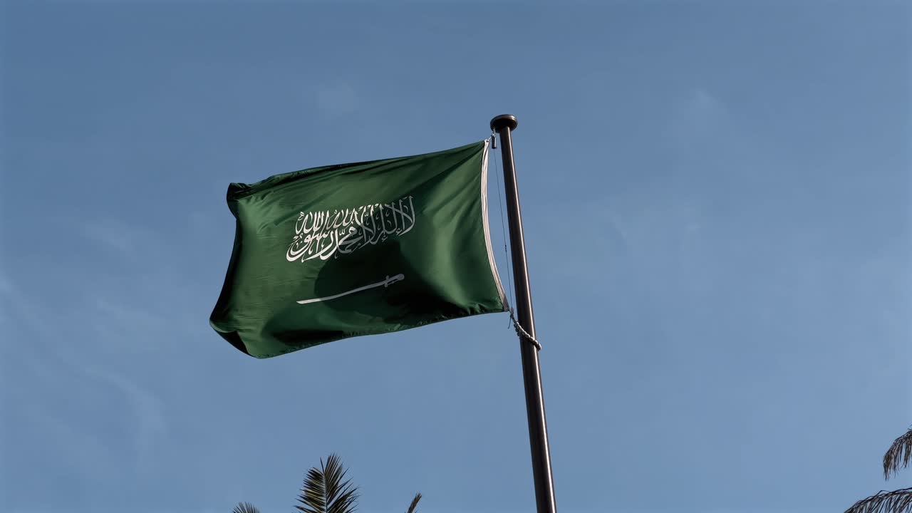 The national flag of Saudi Arabia flutters gracefully on a sunny, cloudless day, with the tips of palm trees swaying gently in the background, symbolizing pride and heritage