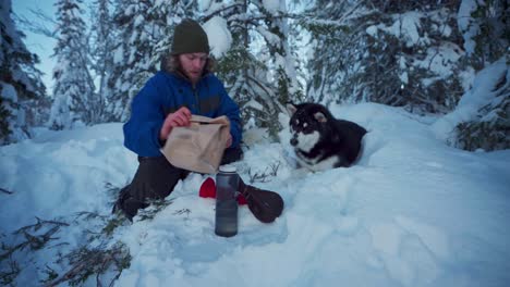 Hombre-Con-Su-Perro-Malamute-De-Alaska-Sentado-En-Un-Suelo-Nevado-Y-Tomando-Un-Refrigerio-Durante-El-Descanso-En-Trondheim,-Noruega---Tiro-Estático