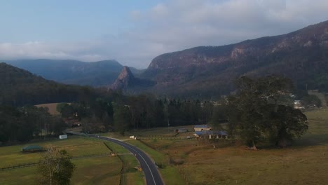 Aerial-view-of-a-country-road-winding-through-a-hinterland-bush-valley-with-a-pinnacle-shaped-mountain-formation-set-against-a-clouded-skyline