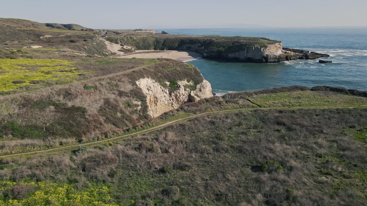 A slow motion drone shot flying right over yellow California poppies then tilting down to show turquoise Pacific ocean waves crashing on a small beach north of Santa Cruz off of the historic Highway 1
