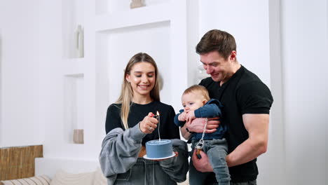 happy caucasian parents celebrating ten months of their child's life. woman is lighting a candle on the cake and man is holding a baby.
