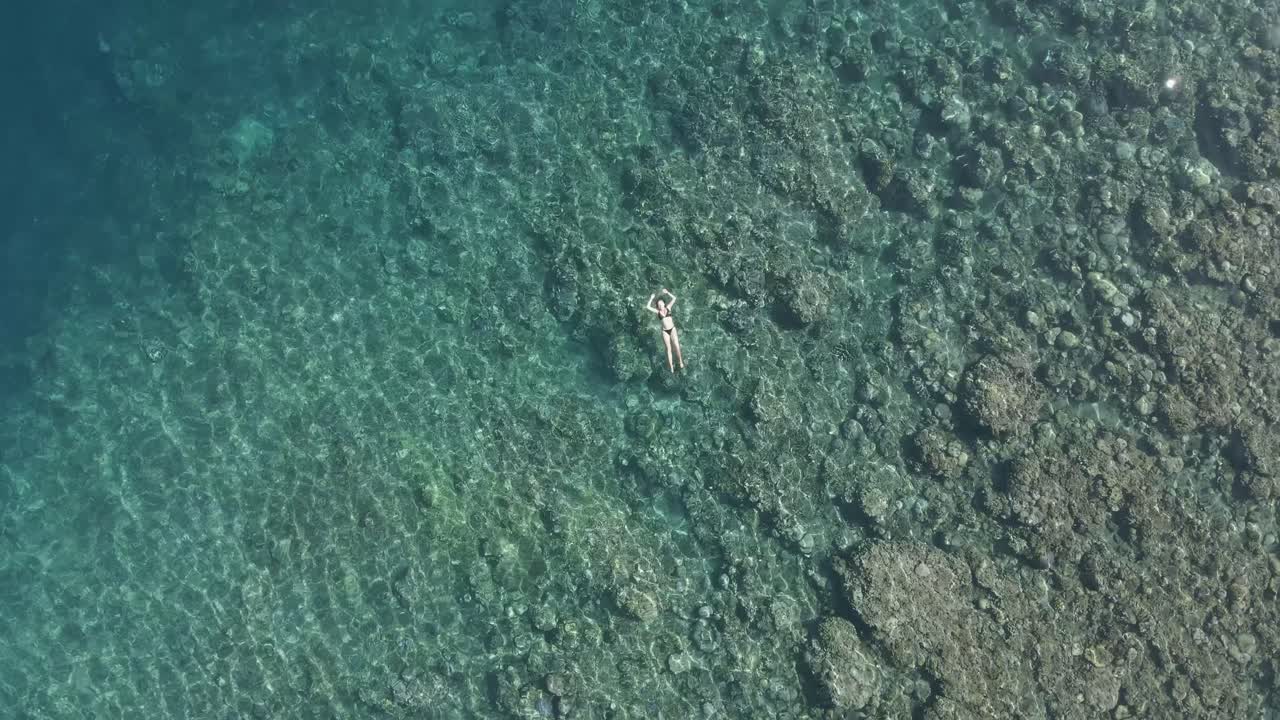 Rotating aerial rises over bikini woman floating in clear shallow reef