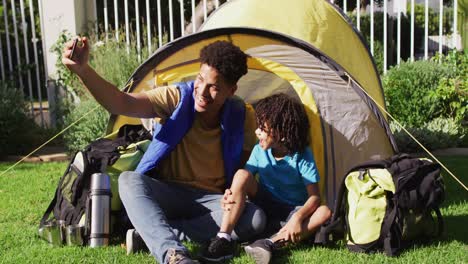 Happy-biracial-man-and-his-son-taking-selfies-using-smartphone-in-garden