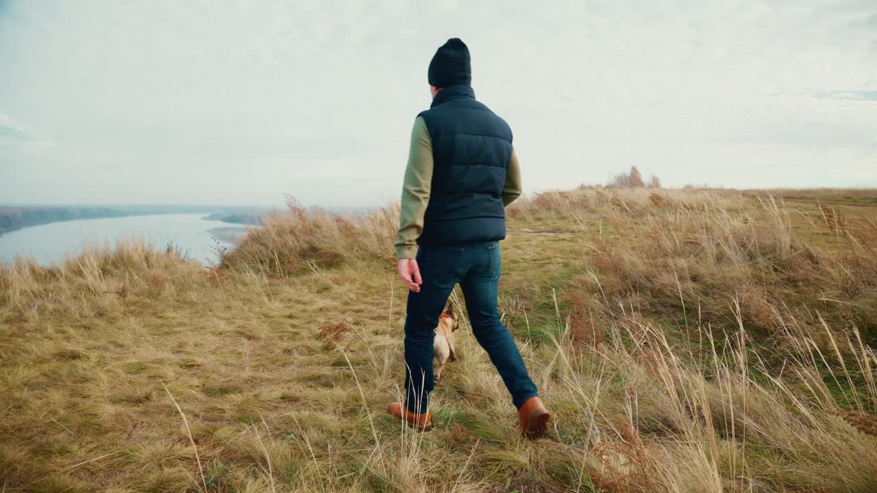 Adventurous Stroll: A Person Enjoys a Peaceful Walk with Their Dog Along the Scenic Meadow Overlooking the Serene Water Body, Embracing Nature's Beauty and Bonding Moments