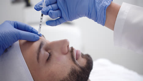beautiful slow motion shot of a female doctor's hand in blue gloves injecting botox into the forehead of a young bearded male patient lying on a stretcher.