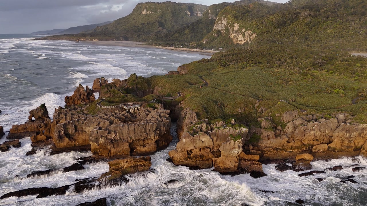 Panoramic aerial view of Pancake Rocks in Punakaiki on West Coast of New Zealand’s South Island at sunset, waves crashing against rugged cliffs under warm golden light. Holiday trip.