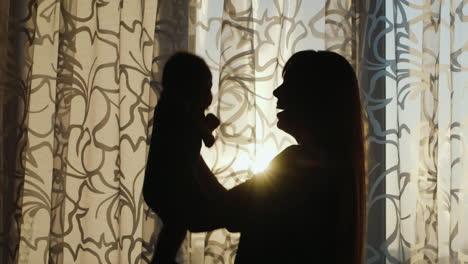 Silhouette-Of-A-Young-Mother-Playing-With-Baby-By-The-Window-At-Sunset-Happy-Childhood