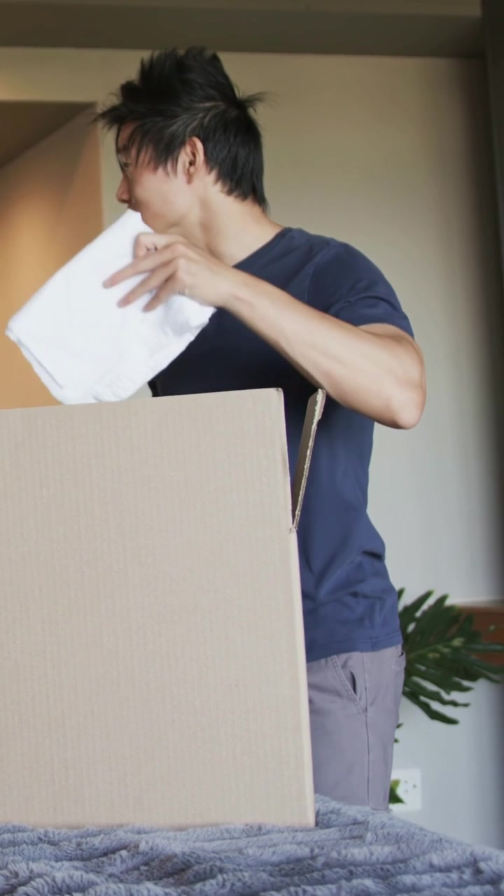Video vertical: Hombre con camiseta azul marino tirando de un paquete blanco, metiendo la mano en una caja en casa después de escuchar una voz.