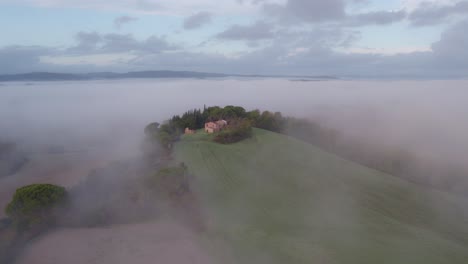 con la casa de la granja de la colina que sobresale de la niebla en italia, aérea