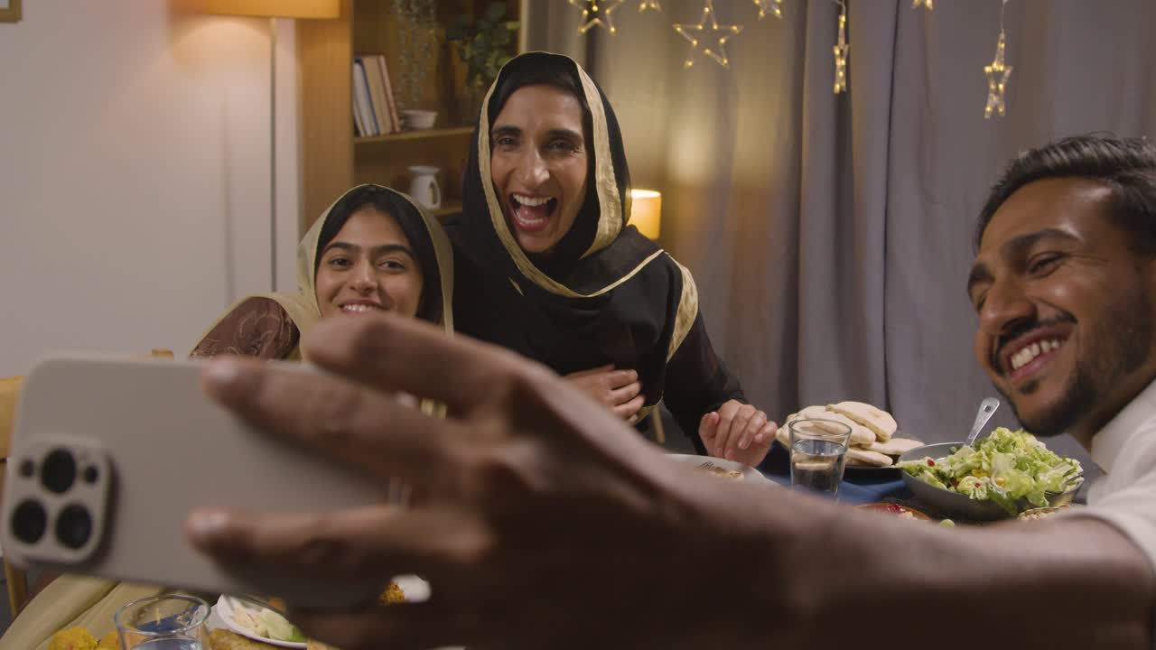 familia musulmana sentada alrededor de una mesa en casa con comida para comer celebrando eid posando para selfie 1