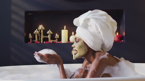 African-american-woman-with-towel-and-mask-taking-bath-and-blowing-foam-in-bathroom