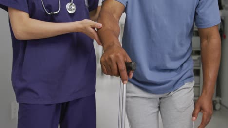 sección media de una doctora diversa que apoya al paciente masculino en el uso del bastón en el hospital