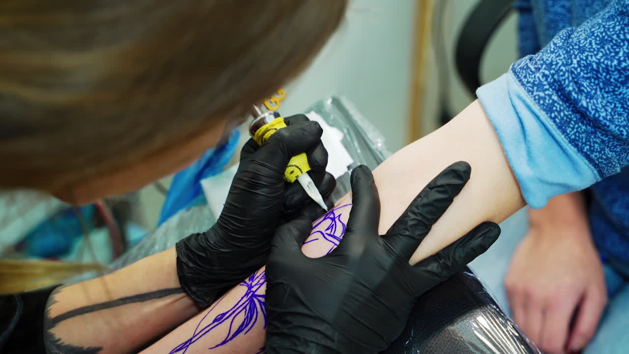 Female tattoo artist carefully using ink machine on the customer's hand. The master works by the blue lines. Close up.