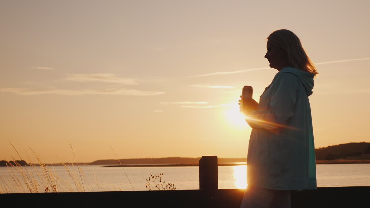 외로운 여자는 일몰을 바라보며 울타리 옆에 홀로 서 있는 호수 위 캔에서 맥주를 마신다