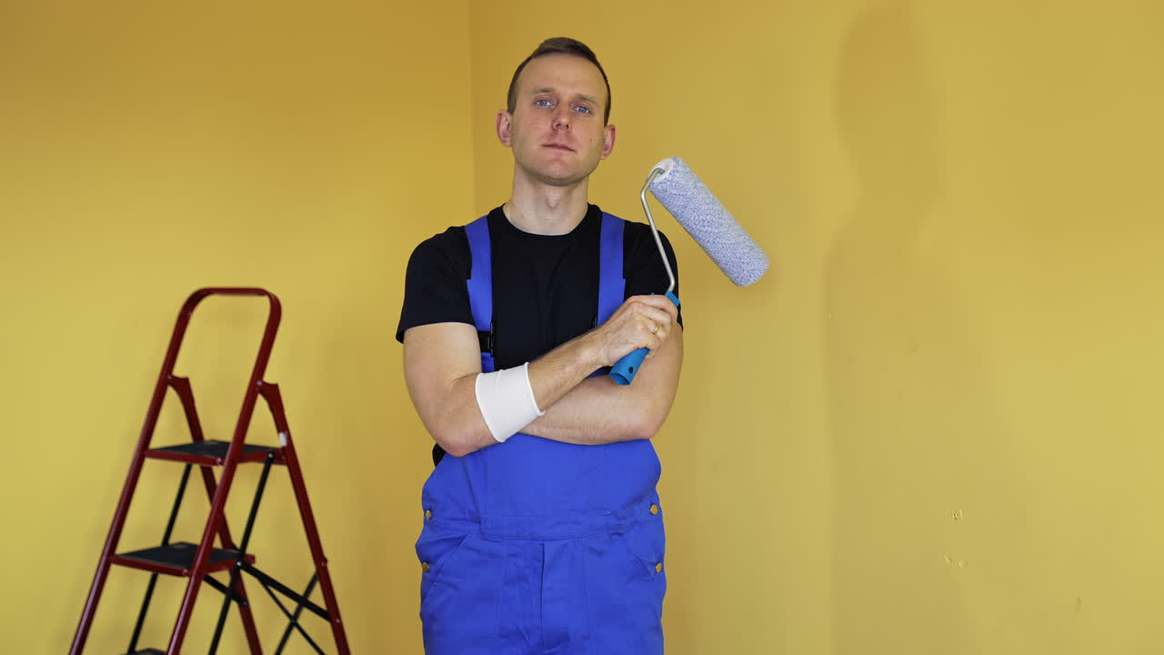 Portrait of a worker painter. Young man in blue overalls standing in the room and holding roller brush before doing a makeover in flat. Home improvement and renovation.