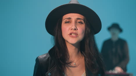 close-up of a lady wearing a black hat and black leather jacket, singing passionately with her hand raised. a blurred keyboardist is visible in the background against a blue backdrop