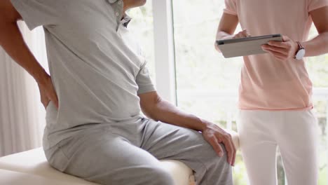 diverse male physiotherapist with tablet examinig senior male patient, in slow motion