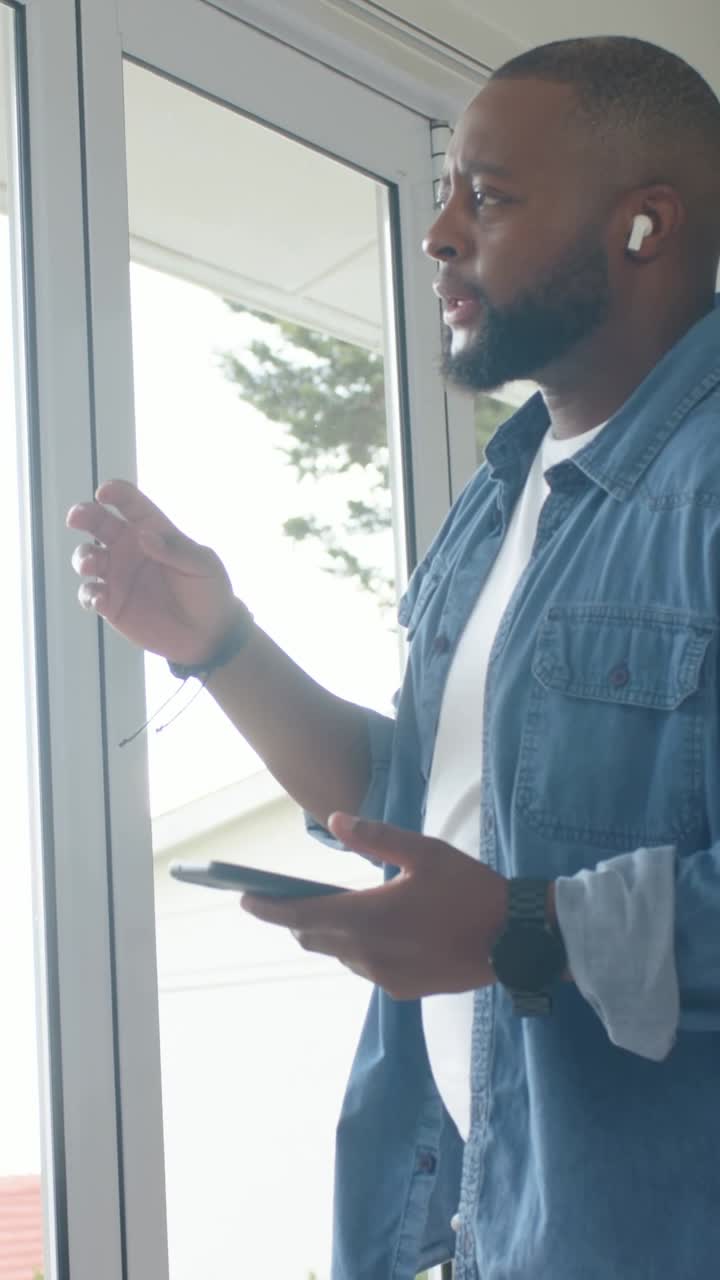 Vertical video: Answering call man in denim holding phone speaking at glass door, earbuds, watch