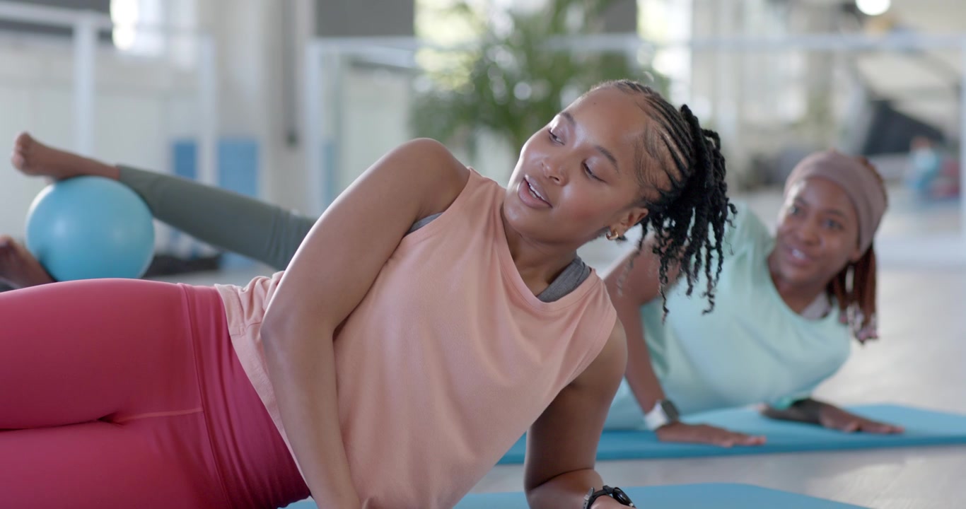 Two women doing side-lying-exercise on blue mats in sunlit studio, lead cueing partner to keep form