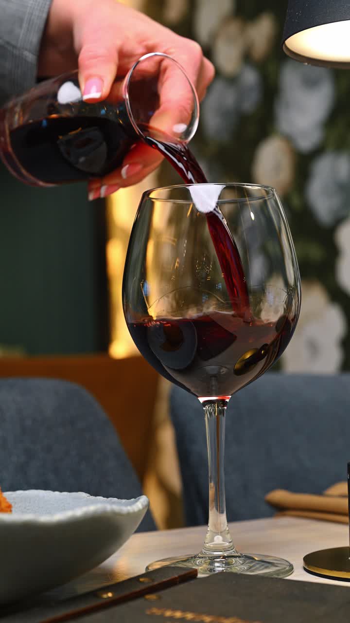 Vino rosso versato in un grande bicchiere al ristorante. Primo piano di vino rosso scuro che viene versato da un decanter in un bicchiere trasparente con movimento.