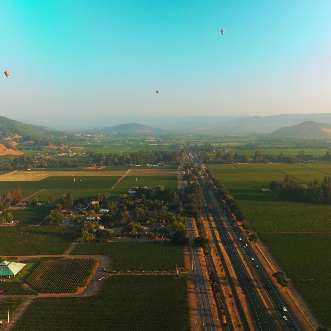 Stunning beautiful green valley with some vineyards. Hot air balloons in blue sky over the land. Hazy mountains at backdrop