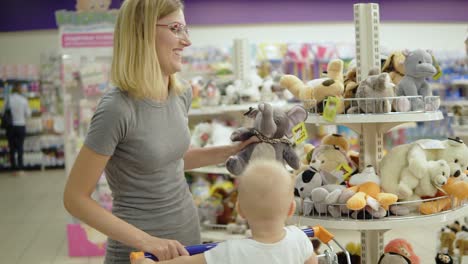 smiling mother showing her child a toy elephant in a toy section for children in the supermarket while her cute little child