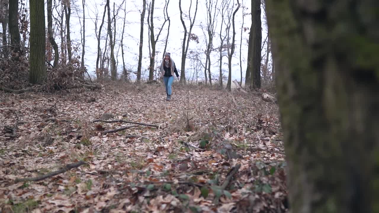 Revealing shot of young smiling caucasian girl with hat and scarf walking into forest during cold winter day, SLOW MOTION