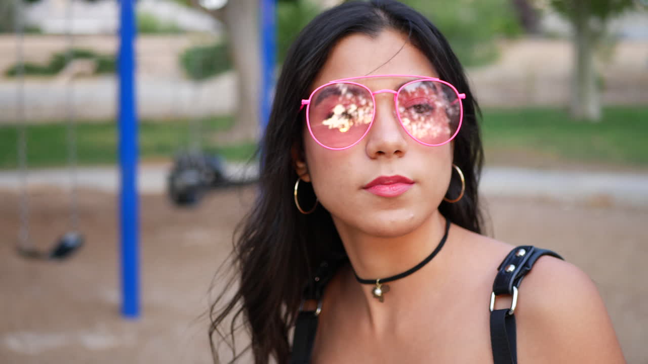 An attractive hispanic woman hipster with retro pink sunglasses in a colorful park playground swing set SLOW MOTION
