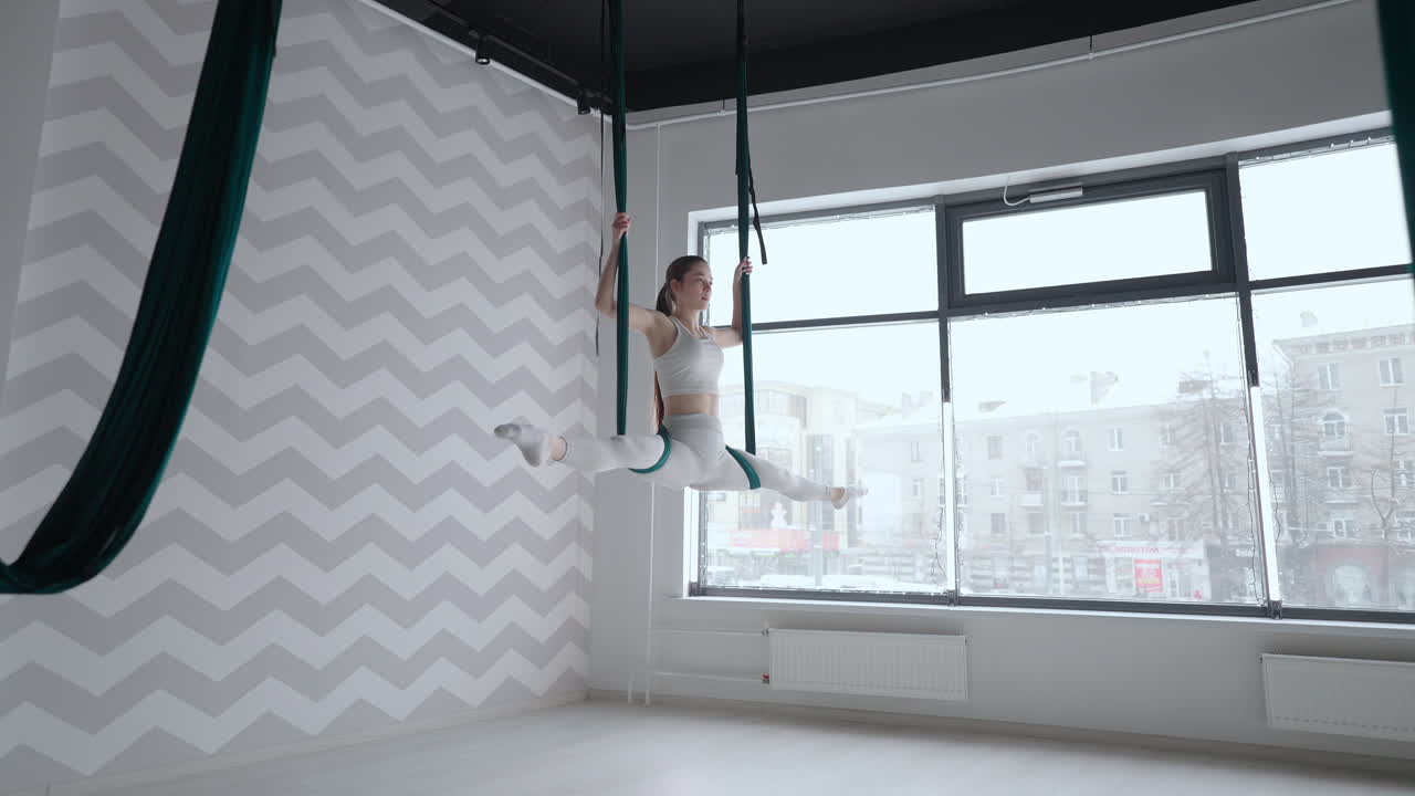 молодая красивая женщина выполняет упражнения на гамаке из аэро-йоги. женщина днем перед зеркалом в зале выполняет растяжку в одиночку
