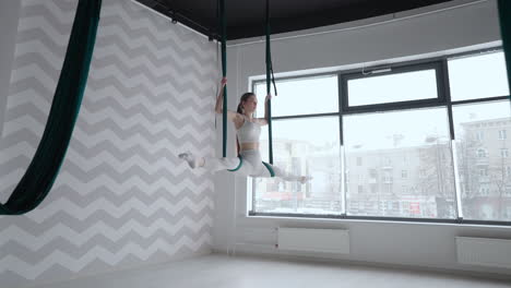 a young beautiful woman performs exercises on a hammock from aero yoga. a woman in the afternoon in front of the mirror in the hall performs stretching alone