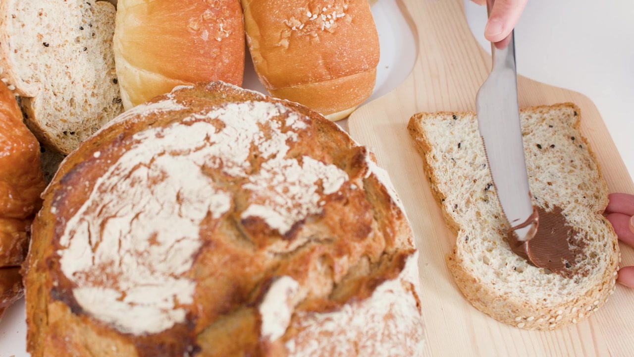 A knife spreads chocolate hazelnut cream on sliced bread amidst various fresh bakery items.