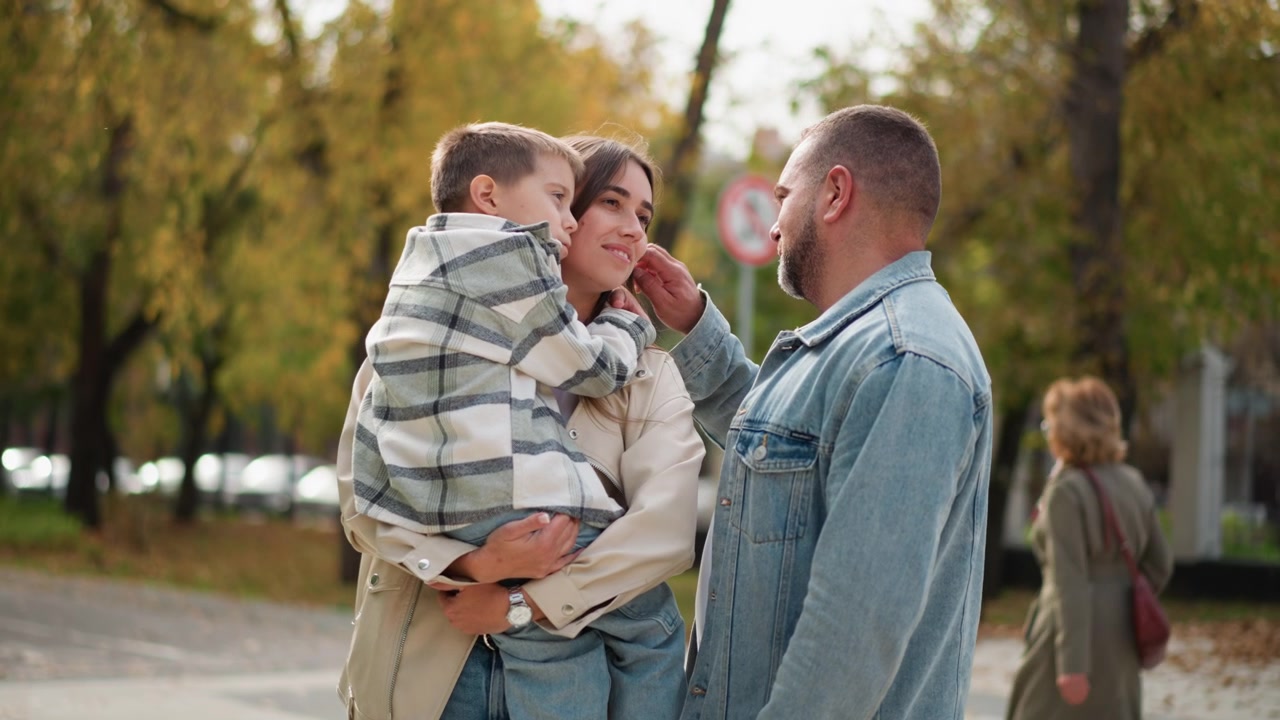 Primo piano di una famiglia all'aperto con l'uomo che accarezza delicatamente i capelli del figlio mentre sono in piedi insieme in una giornata autunnale. La donna tiene in braccio il loro figlio piccolo e sono circondati da alberi con colori autunnali.