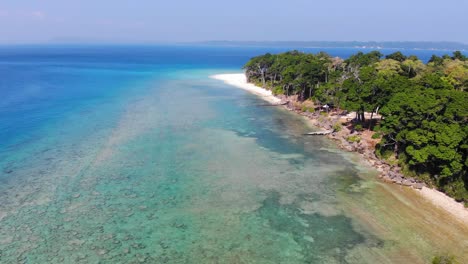 drone de izquierda a derecha que muestra islas remotas de andaman en el océano azul a lo lejos con un bosque antiguo que bordea una playa y arrecifes y rocas para ser vistos bajo las aguas poco profundas
