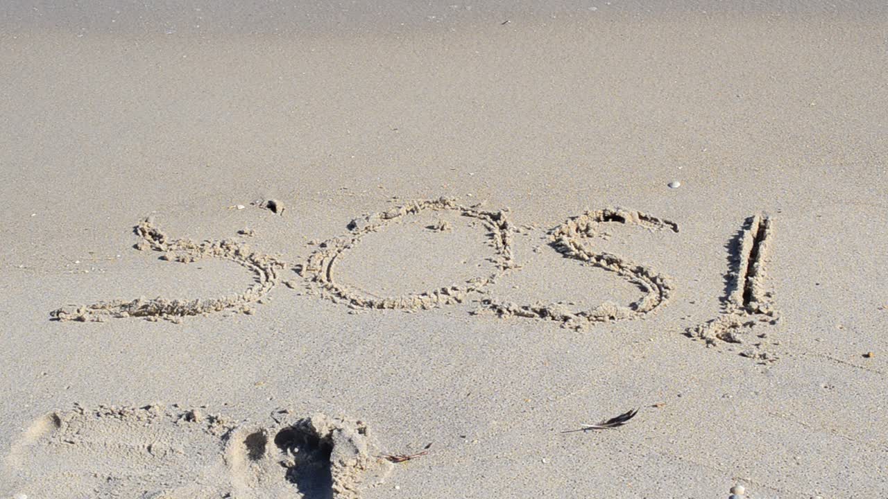 Et råb om hjælp på sandstranden. SOS-tegnet skylles væk af en bølge.
