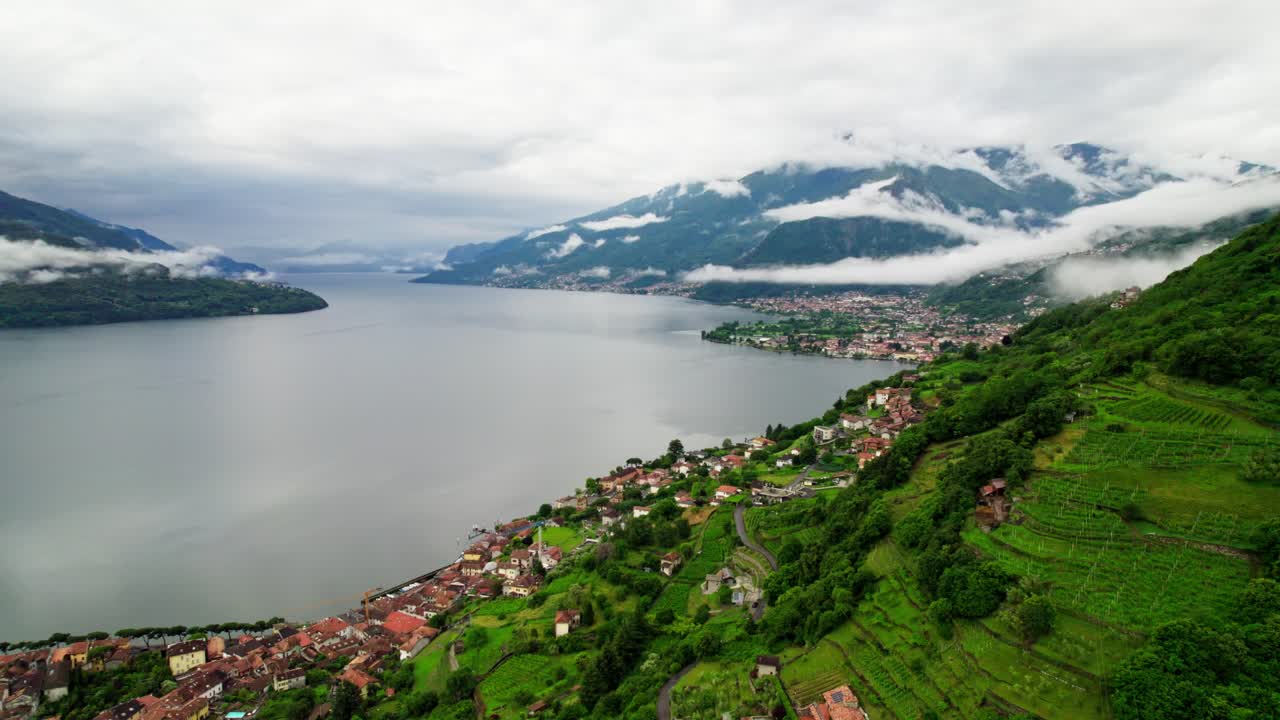 Filmato aereo cinematografico realizzato con drone che sorvola verso sinistra una città storica con tetti rossi, il lago di Como e montagne nuvolose sullo sfondo