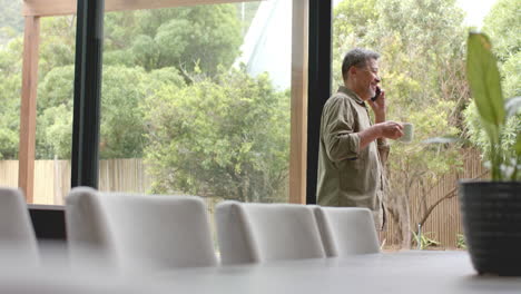happy senior biracial man by window talking on smartphone holding coffee, copy space, slow motion