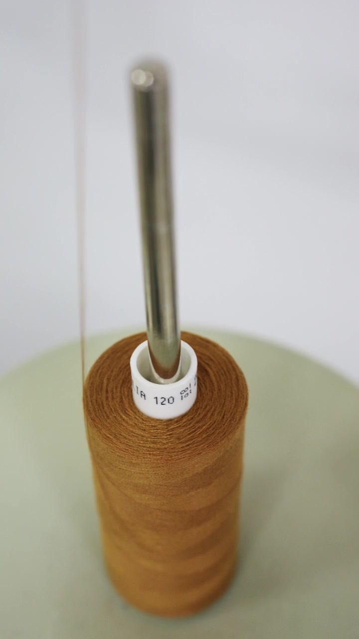 Close up of a mustard yellow thread cone on a thread stand, with a fine strand stretching across a white studio background