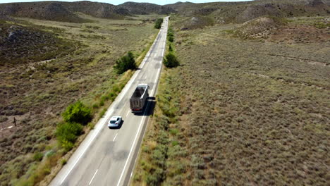 Luftaufnahme-Eines-Autos-Auf-Der-Straße,-Das-Einen-Lastwagen-überholt,-Der-An-Einem-Wüstenort-Entlang-Fährt,-Mit-Windmühlen-Im-Hintergrund