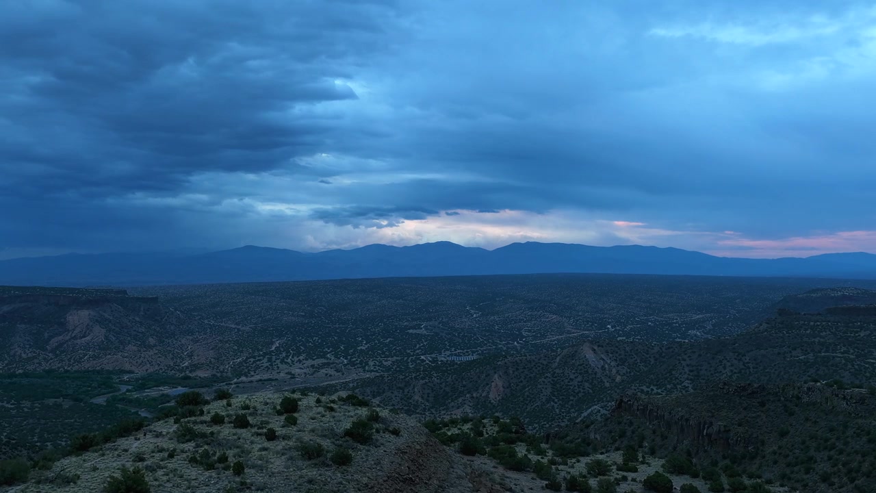 4K UHD drone-timelapse, der sporer over en barsk mesa-kant, mens dybblå skumring falder på. Tunge stormskyer bevæger sig hen over den store ørkendal mod en fjern sydvestlig bjergkæde i horisonten