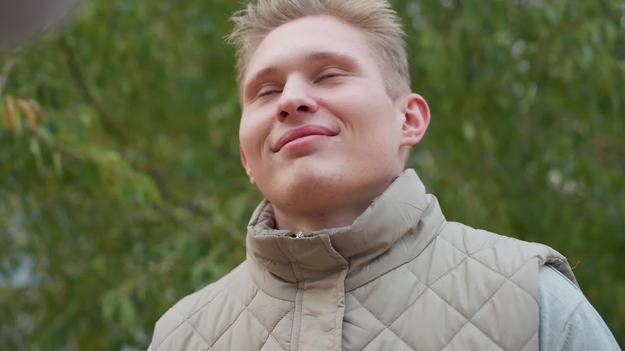 Young man kneels with warm genuine smile during emotional moment surrounded by swaying green leaves in soft breeze while wearing light quilted jacket