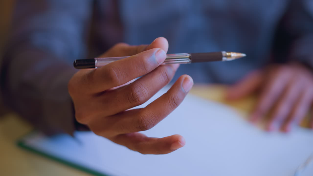 Close-up of male hand holding pen over clipboard, suggesting pause before writing or decision-making moment during discussion or interview, with focused gesture conveying readiness, contemplation, or intention in indoor workspace