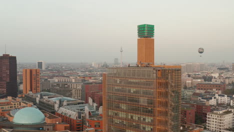 Pull-back-footage-of-man-standing-on-roof-of-tall-building-in-modern-urban-neighbourhood.-Hot-air-balloon-and-Fernsehturm-TV-tower-in-distance.-Berlin,-Germany