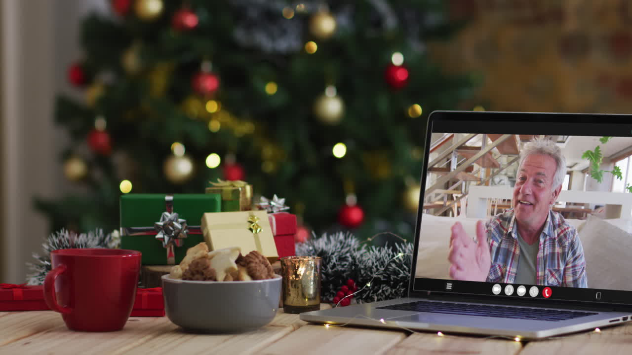 hombre caucásico de alto nivel saludando en una videollamada en una computadora portátil, con decoraciones de navidad y árbol