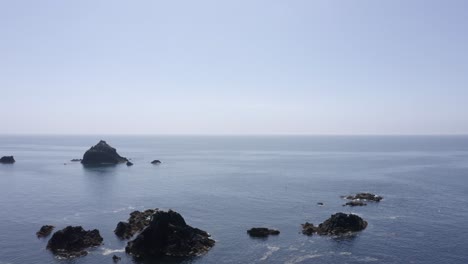 ocean aerial flies past jagged rocks toward flat blue water horizon